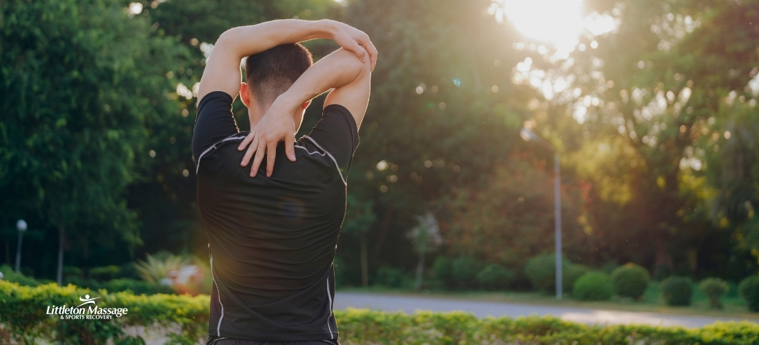 Male athlete stretching shoulders outdoors in a park, supporting injury prevention as part of a sports massage treatment routine.