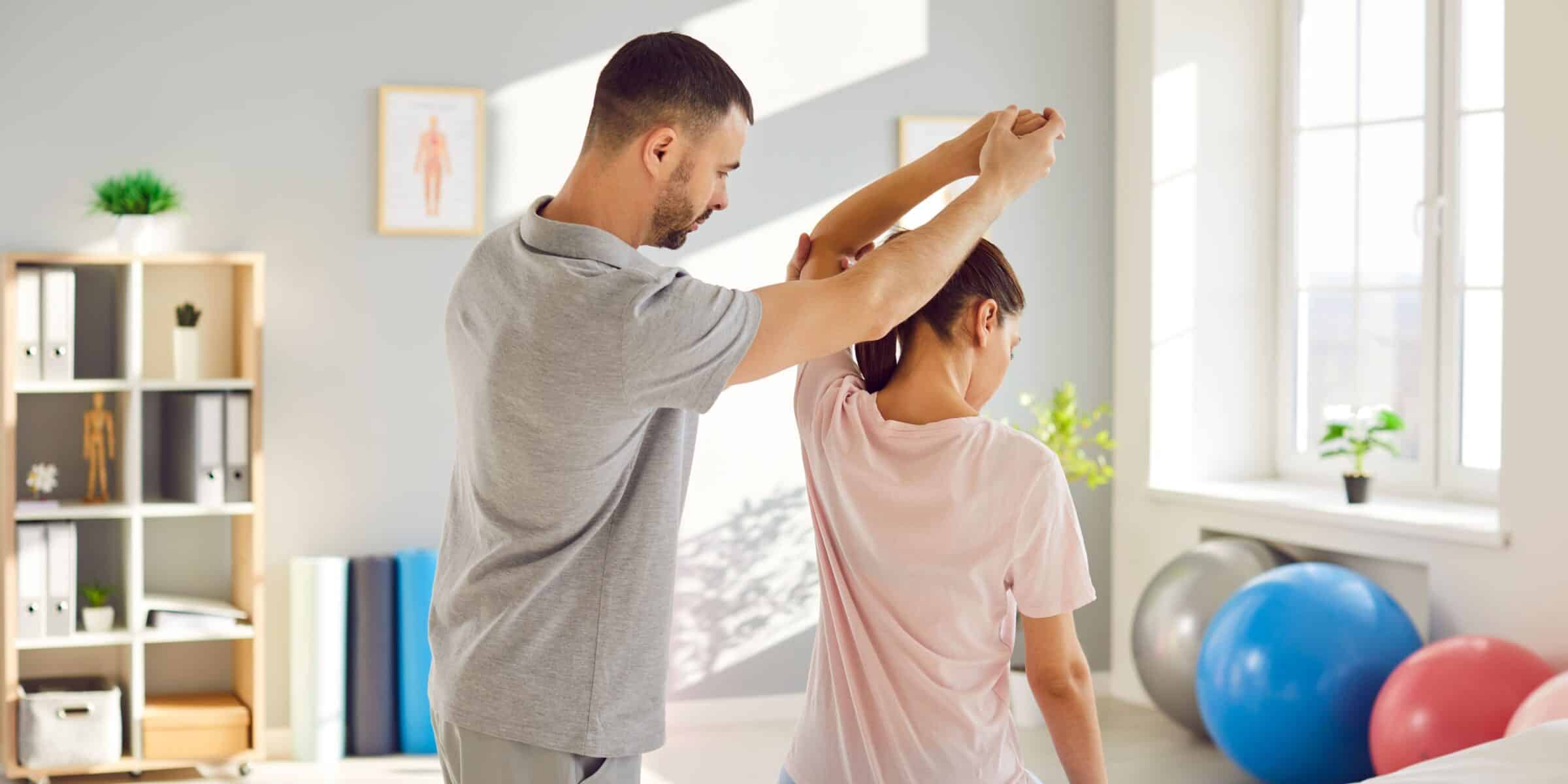 functional sports therapist helping stretch a young woman's arm above her head in a medical office setting