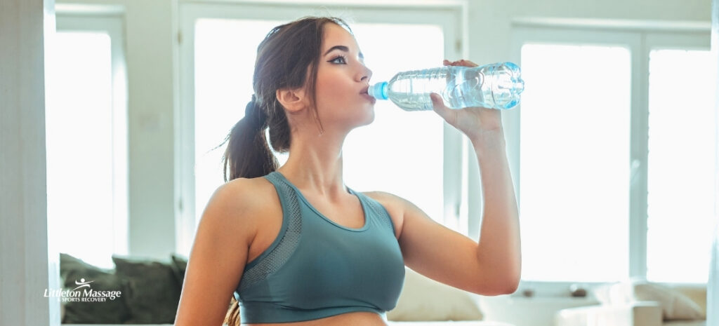 Woman in athletic wear drinking from a water bottle indoors after a massage, illustrating hydration as part of massage aftercare.