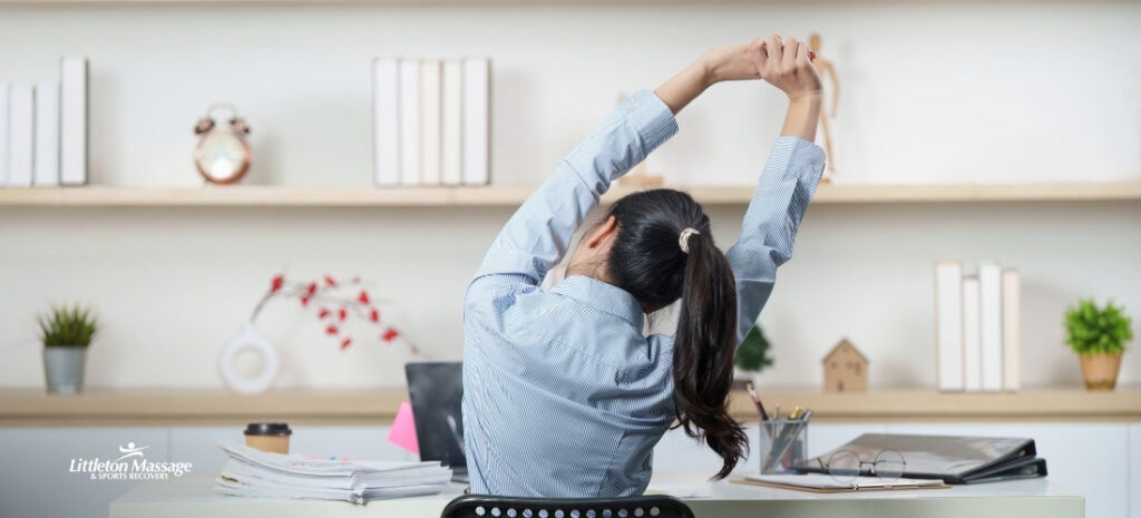 Woman stretching arms overhead at her desk, highlighting the need for assisted stretching to relieve tension